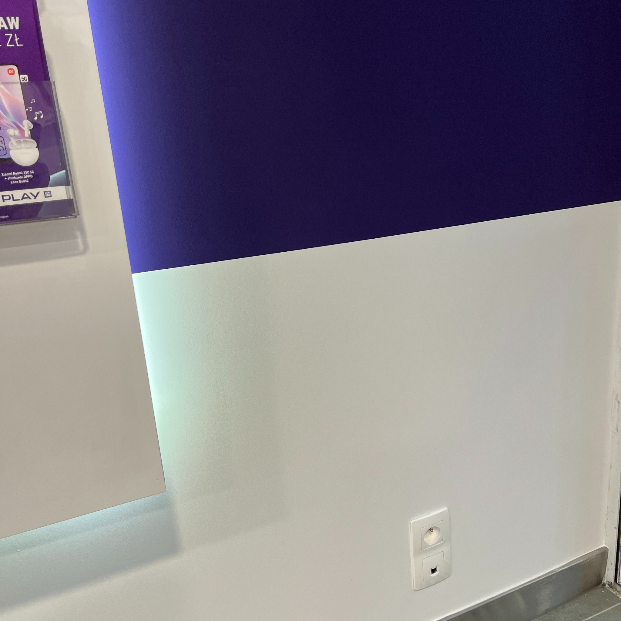 Ściana pomalowana na biało i fioletowo z gniazdkiem elektrycznym i fragmentem plakatu reklamowego w lewym górnym rogu.
