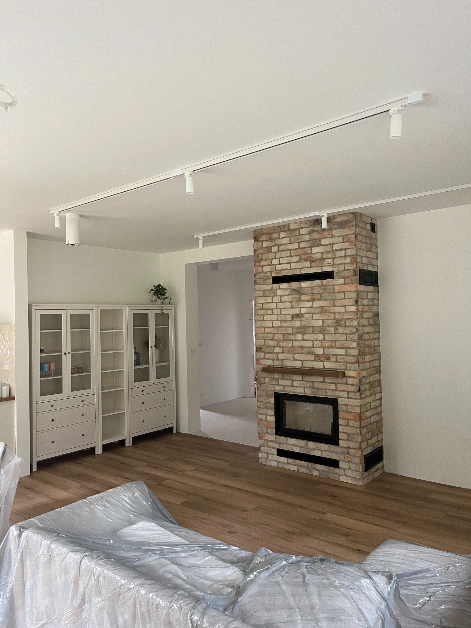 Salon w trakcie wykańczania: ceglany kominek z czarnym wkładem, drewniana podłoga, białe meble i oświetlenie szynowe. Meble zabezpieczone folią malarską.