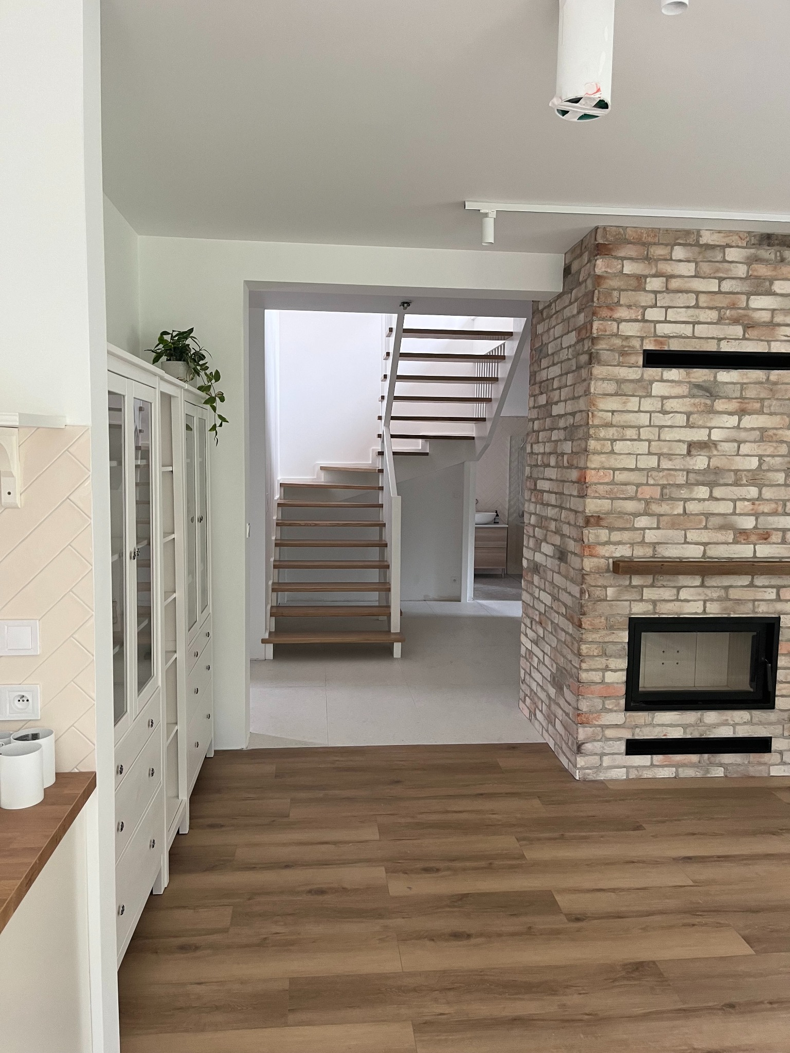 Jasne wnętrze domu z drewnianą podłogą, białymi schodami i ceglanym kominkiem z czarnym wkładem, widok na szafkę z przeszklonymi drzwiami.