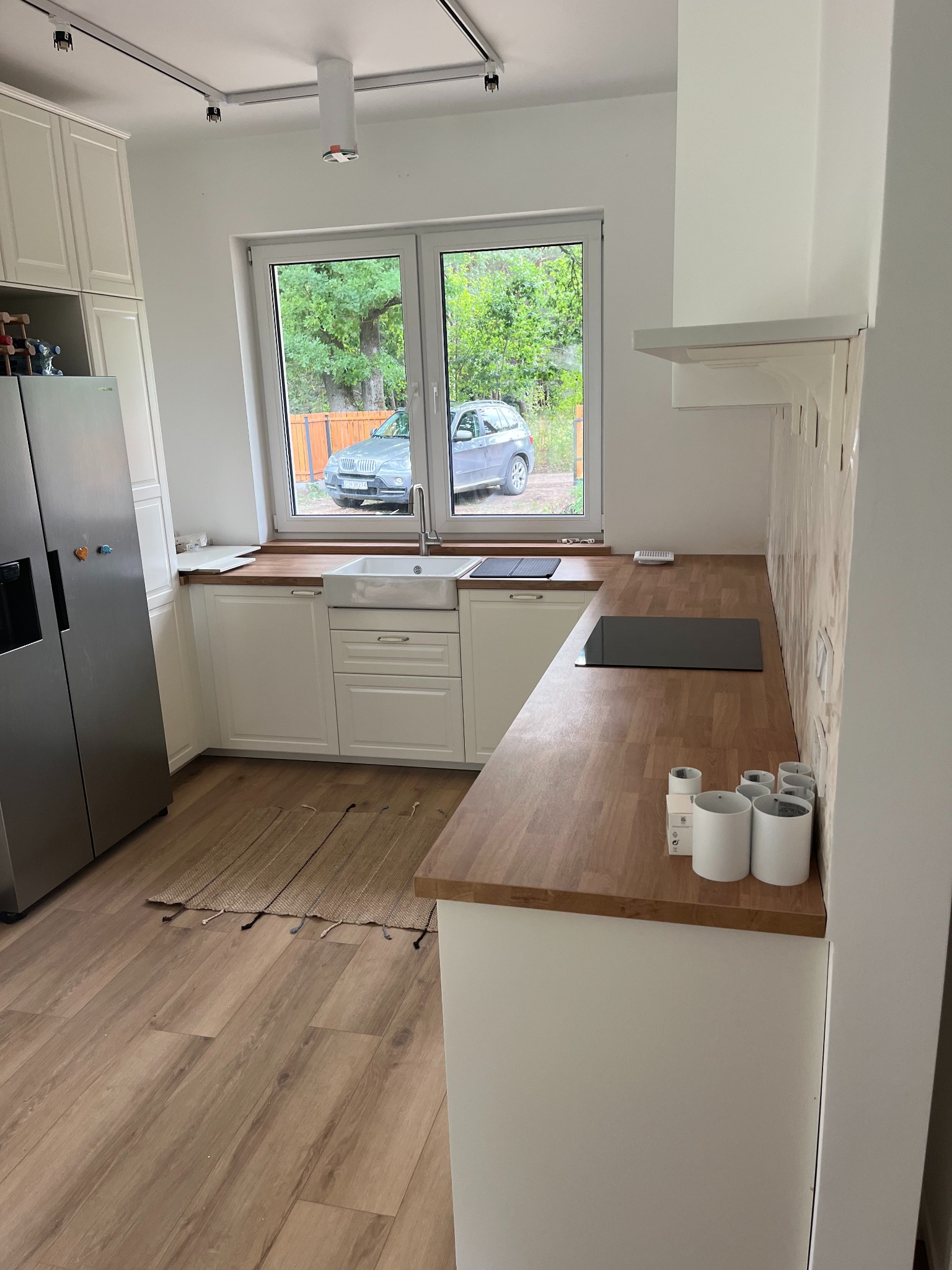 Jasna kuchnia z drewnianym blatem, białymi szafkami, ceramicznym zlewem i widokiem na samochody za oknem.