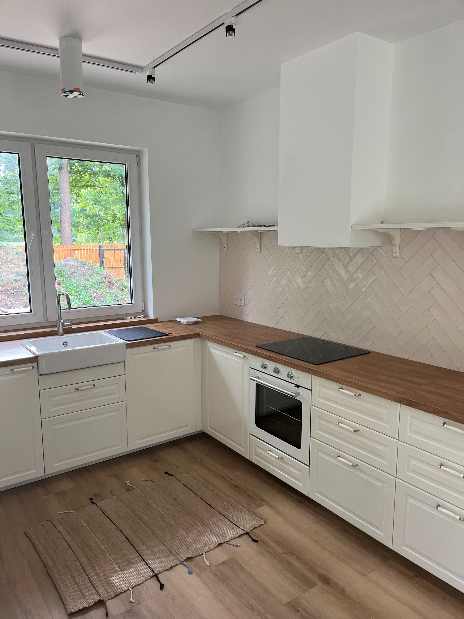 Jasna kuchnia z drewnianym blatem, białymi szafkami z frezowanymi frontami, płytkami ułożonymi w jodełkę na ścianie i jutowym dywanikiem na podłodze.