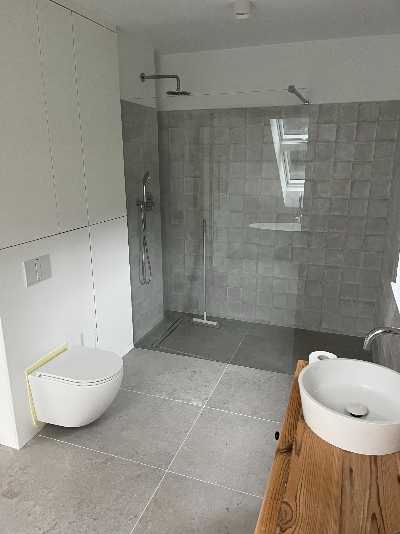 Nowoczesna łazienka w odcieniach szarości, z kabiną prysznicową typu walk-in, podwieszaną toaletą i umywalką nablatową na drewnianym blacie.
