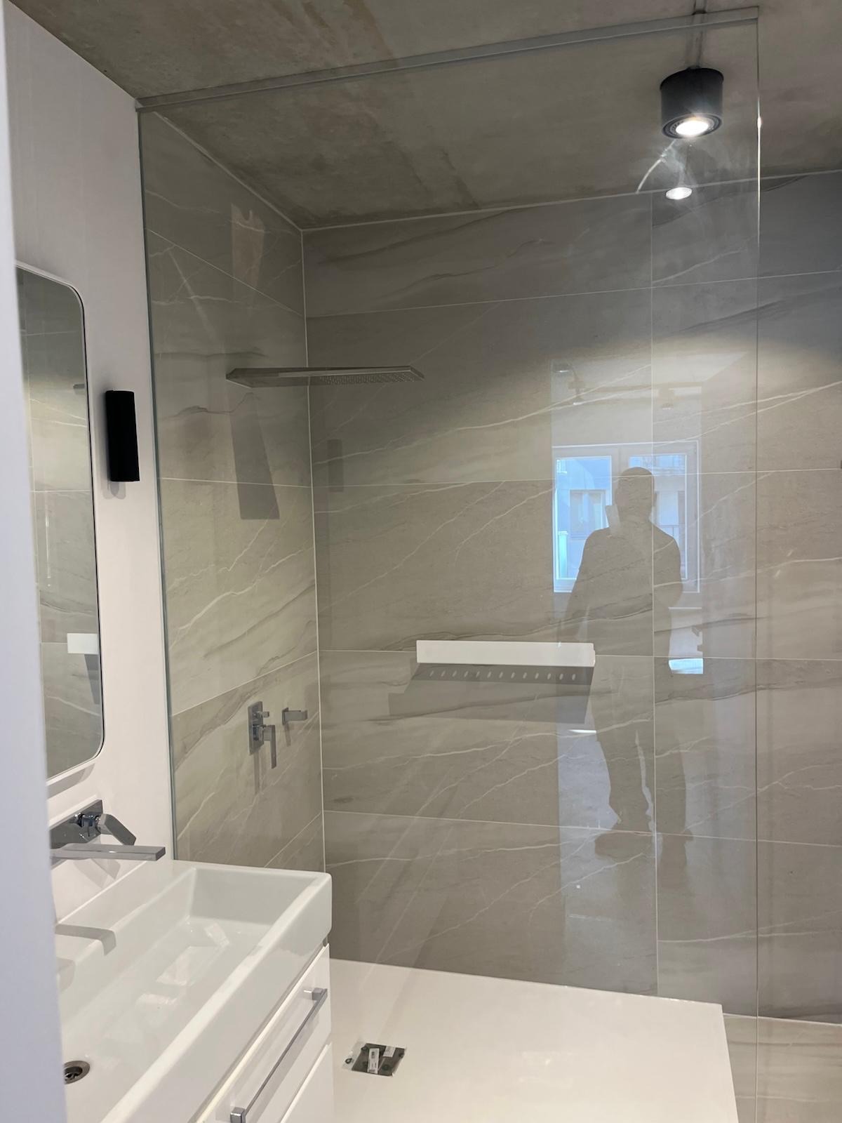 Nowoczesna łazienka z kabiną prysznicową typu walk-in, szare płytki imitujące marmur, biała umywalka nablatowa i minimalistyczna armatura. Widoczne odbicie osoby w szkle.