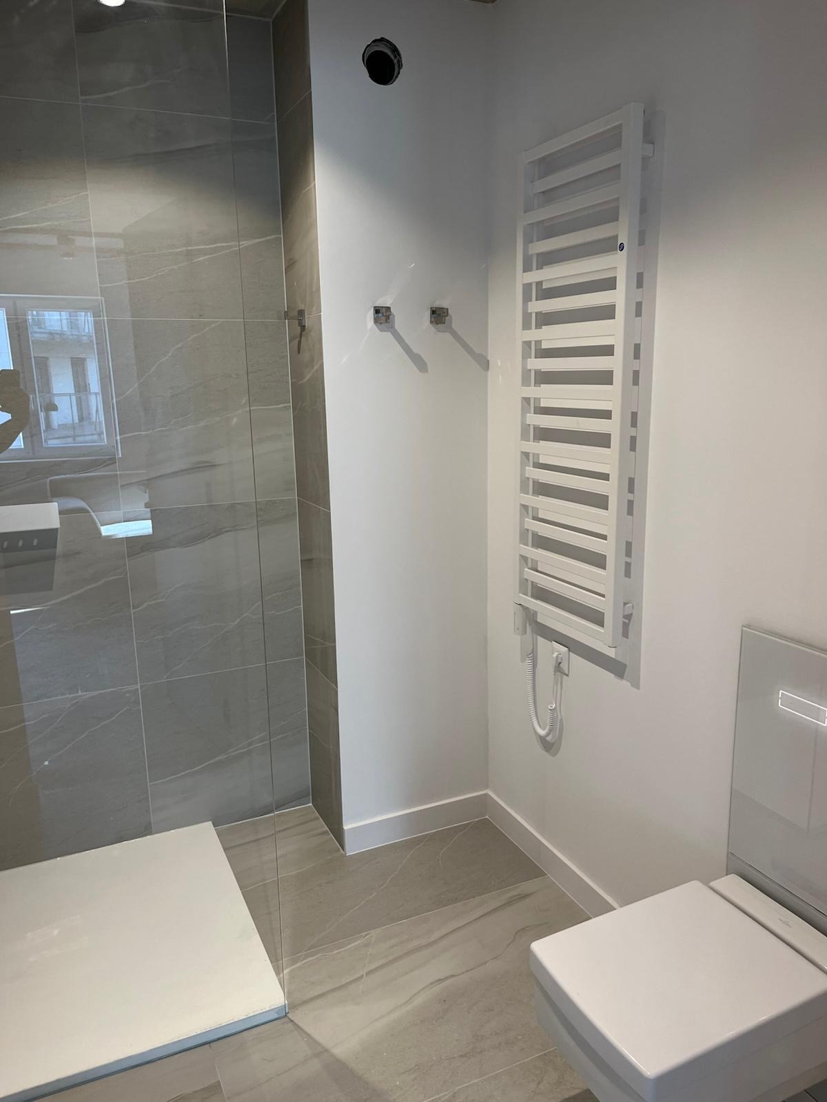 Nowoczesna łazienka po remoncie: szklana kabina prysznicowa z szarymi płytkami imitującymi marmur, biały grzejnik drabinkowy i podwieszana toaleta.