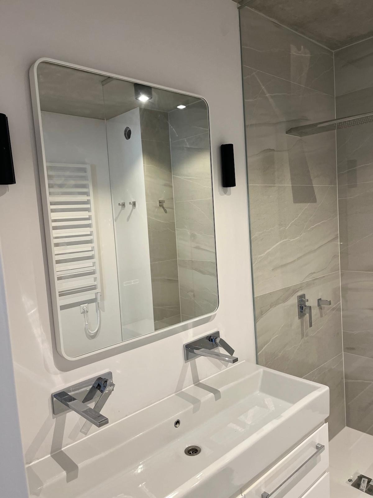 Nowoczesna łazienka z umywalką, lustrem z zaokrąglonymi rogami i szarą marmurową ścianą prysznicową. Minimalistyczne baterie i oświetlenie.