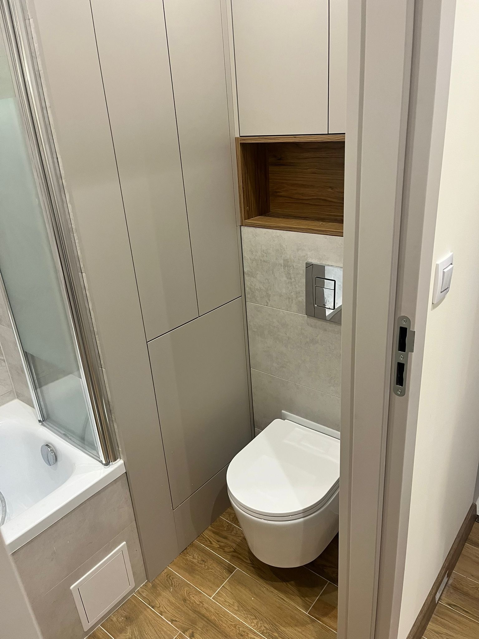 Nowoczesna łazienka z zabudową WC, drewnianą półką i wanną z parawanem. Jasne kolory, drewnopodobna podłoga. Widok zza futryny.