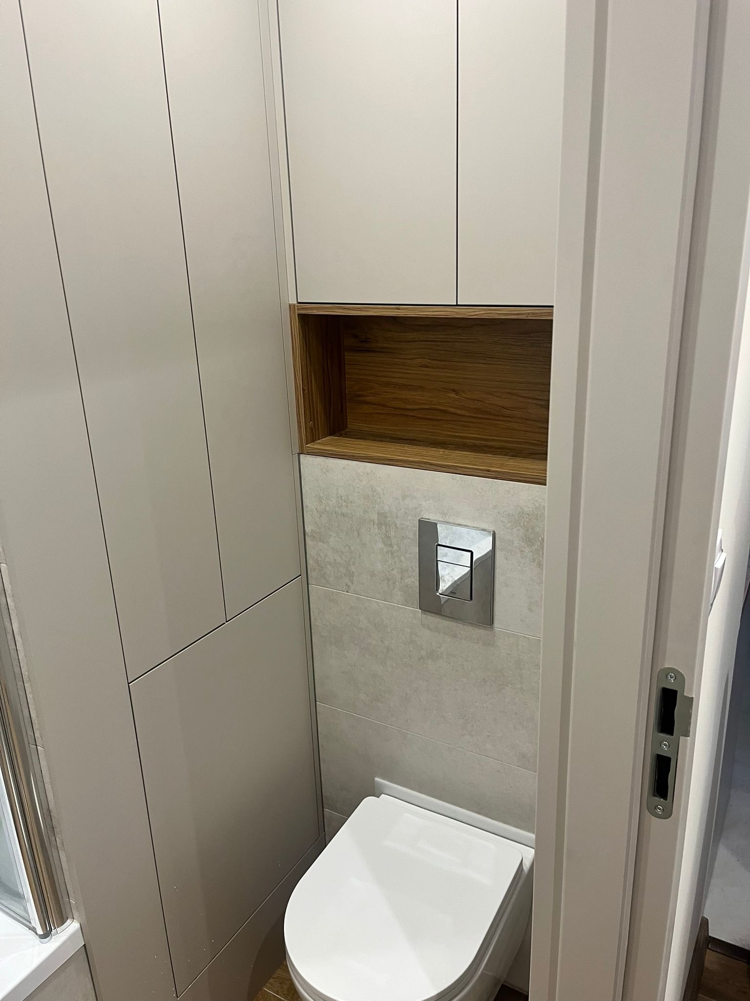 Nowoczesna toaleta z zabudową meblową w odcieniach szarości i drewna. Wisząca miska WC, płytki imitujące kamień, chromowany przycisk spłukujący. Minimalistyczny design.