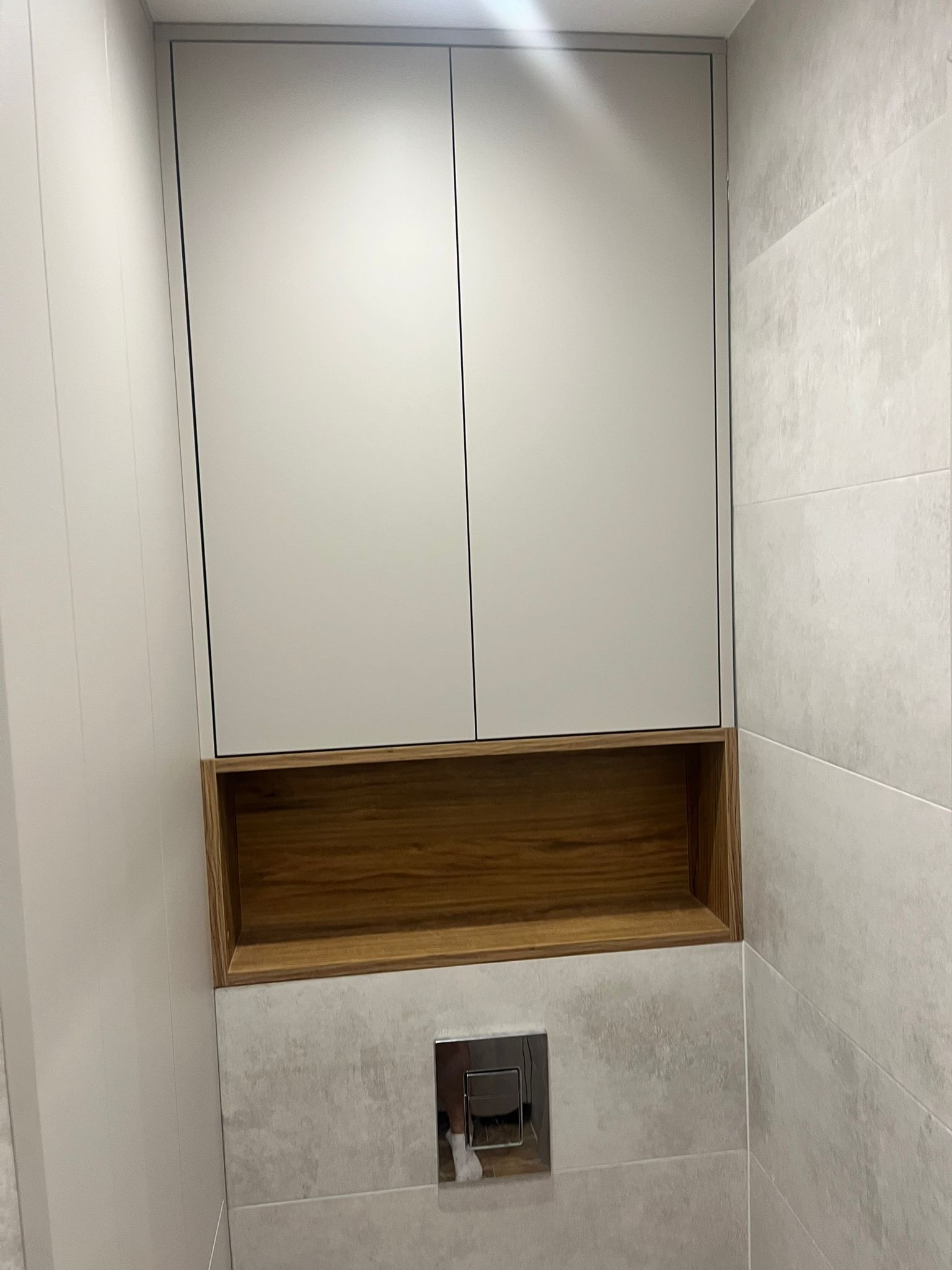 Minimalistyczna zabudowa toalety: białe szafki wiszące z drewnianą wnęką, szare płytki imitujące kamień, chromowany przycisk spłukiwania. Nowoczesne wykończenie łazienki.