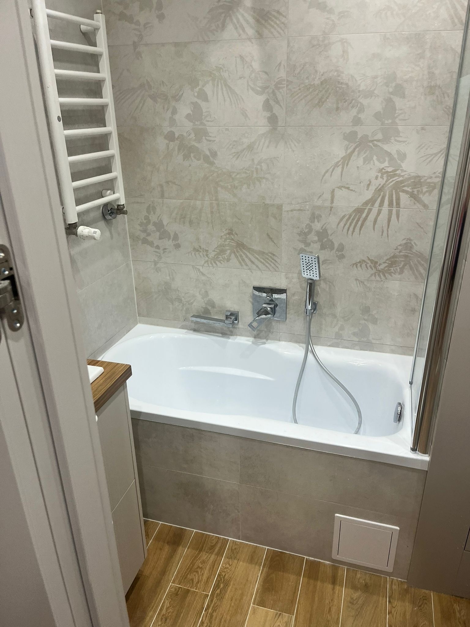 Wykończona łazienka z wanną, baterią, prysznicem i grzejnikiem drabinkowym. Płytki w odcieniach beżu z motywem roślinnym. Widok zza uchylonych drzwi.