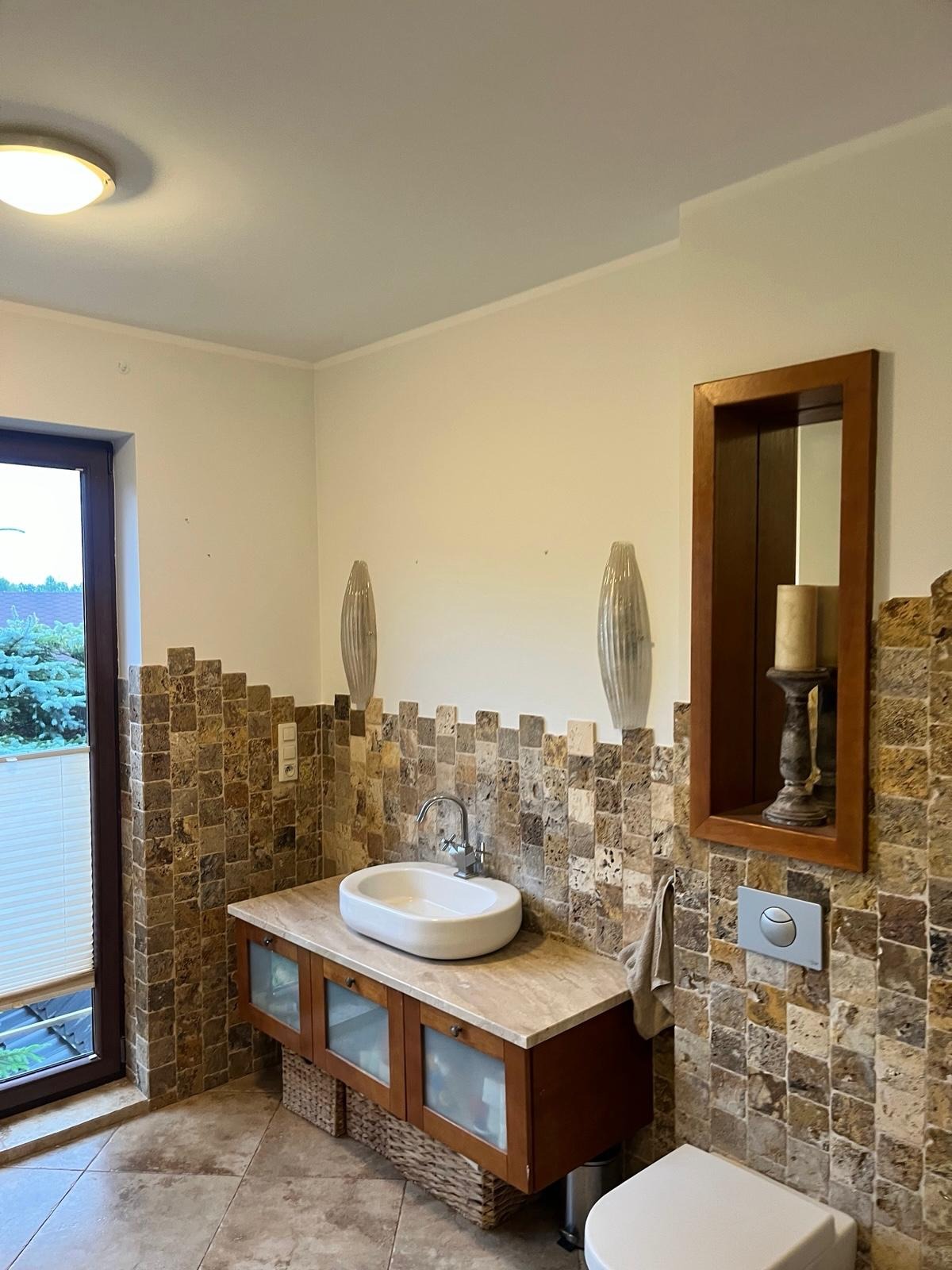 Wykończona łazienka z mozaiką z kamienia naturalnego, umywalką nablatową i drewnianą szafką. Dekoracyjne oświetlenie i lustro w drewnianej ramie dodają elegancji.