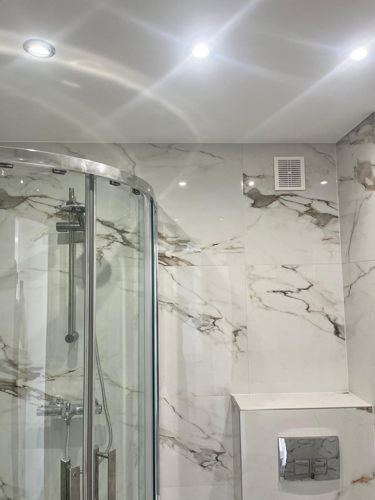 Nowoczesna kabina prysznicowa z chromowanymi elementami i marmurowymi płytkami w odcieniach bieli i szarości. Widoczny fragment wentylacji i przycisk spłuczki.