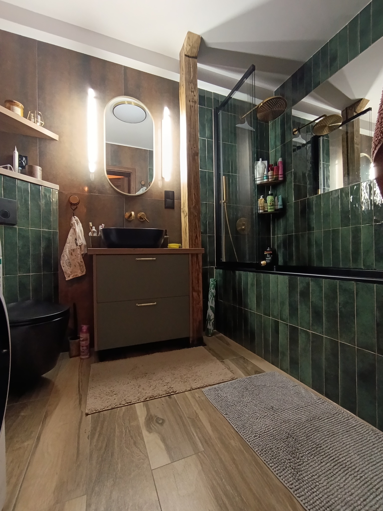 Stylowa łazienka z zielonymi płytkami, drewnianą kolumną i czarną armaturą. Umywalka nablatowa, owalne lustro z podświetleniem i drewniana podłoga.