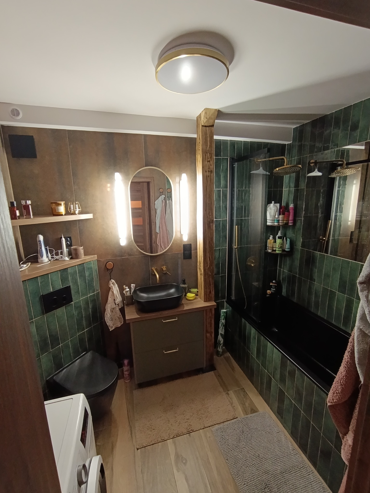 Stylowa łazienka z zielonymi płytkami, czarną armaturą i drewnianymi elementami. Umywalka nablatowa, owalne lustro z podświetleniem, kabina prysznicowa typu walk-in.