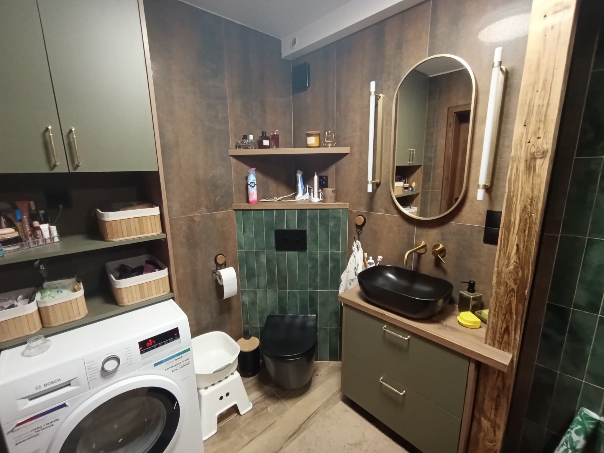 Nowoczesna łazienka w odcieniach zieleni i brązu z czarną armaturą, pralką Bosch i owalnym lustrem. Uwagę przykuwają płytki w stylu retro i drewniane akcenty.