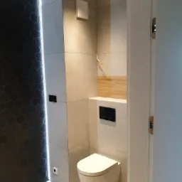 Nowoczesna łazienka z czarną ścianą z heksagonalnych płytek z oświetleniem LED, zawieszona toaleta, szare płytki wielkoformatowe i drewniany akcent nad spłuczką.