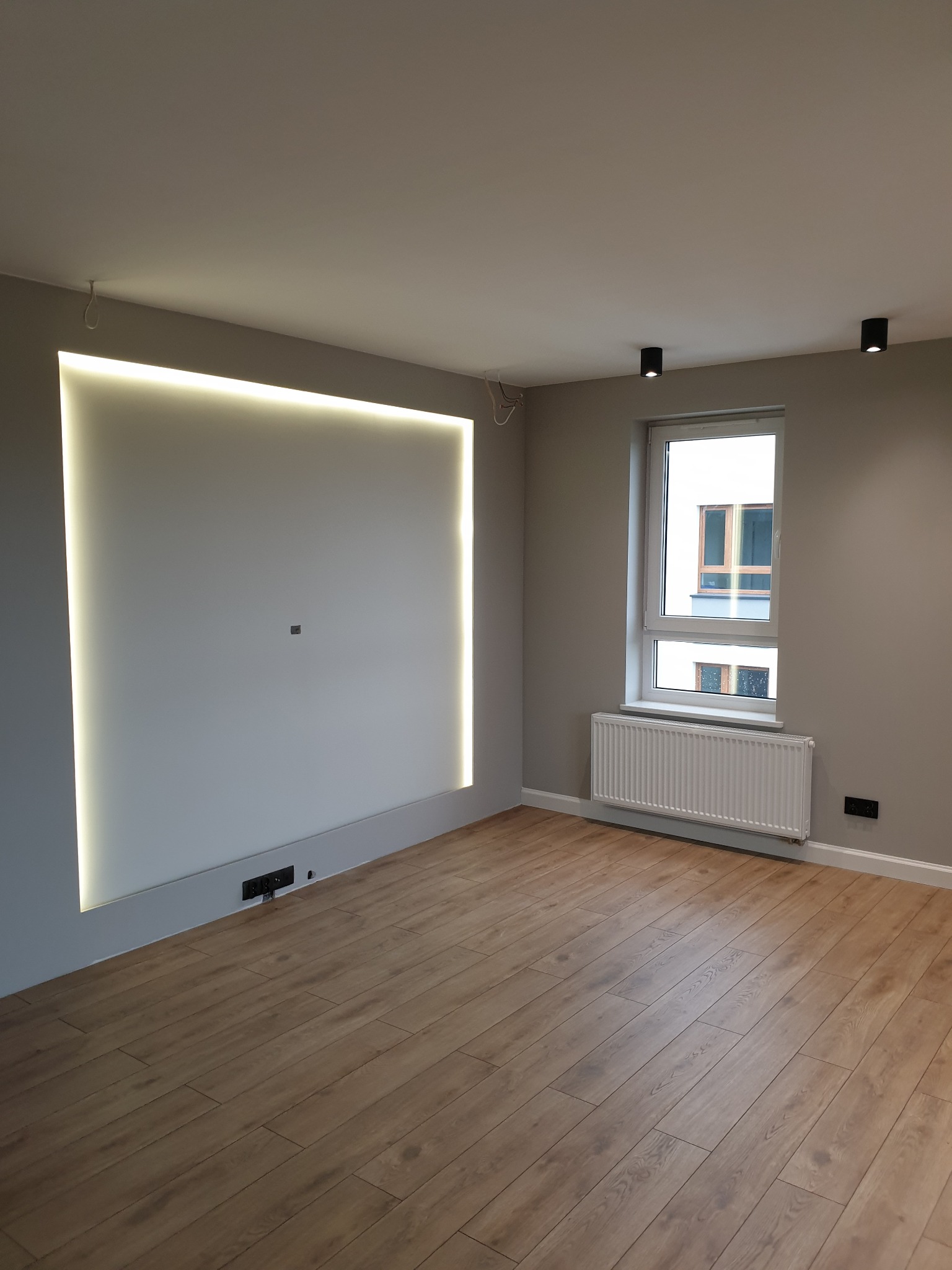 Pusty pokój z drewnianą podłogą, szarą ścianą z podświetlaną wnęką, grzejnikiem pod oknem i dwoma czarnymi lampami sufitowymi.