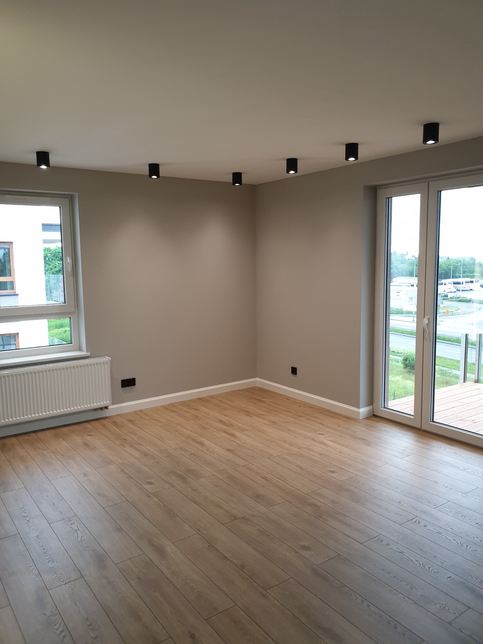 Pusty pokój po remoncie z panelami podłogowymi w odcieniach beżu, szare ściany, białe okna i drzwi balkonowe oraz czarne, punktowe oświetlenie sufitowe.
