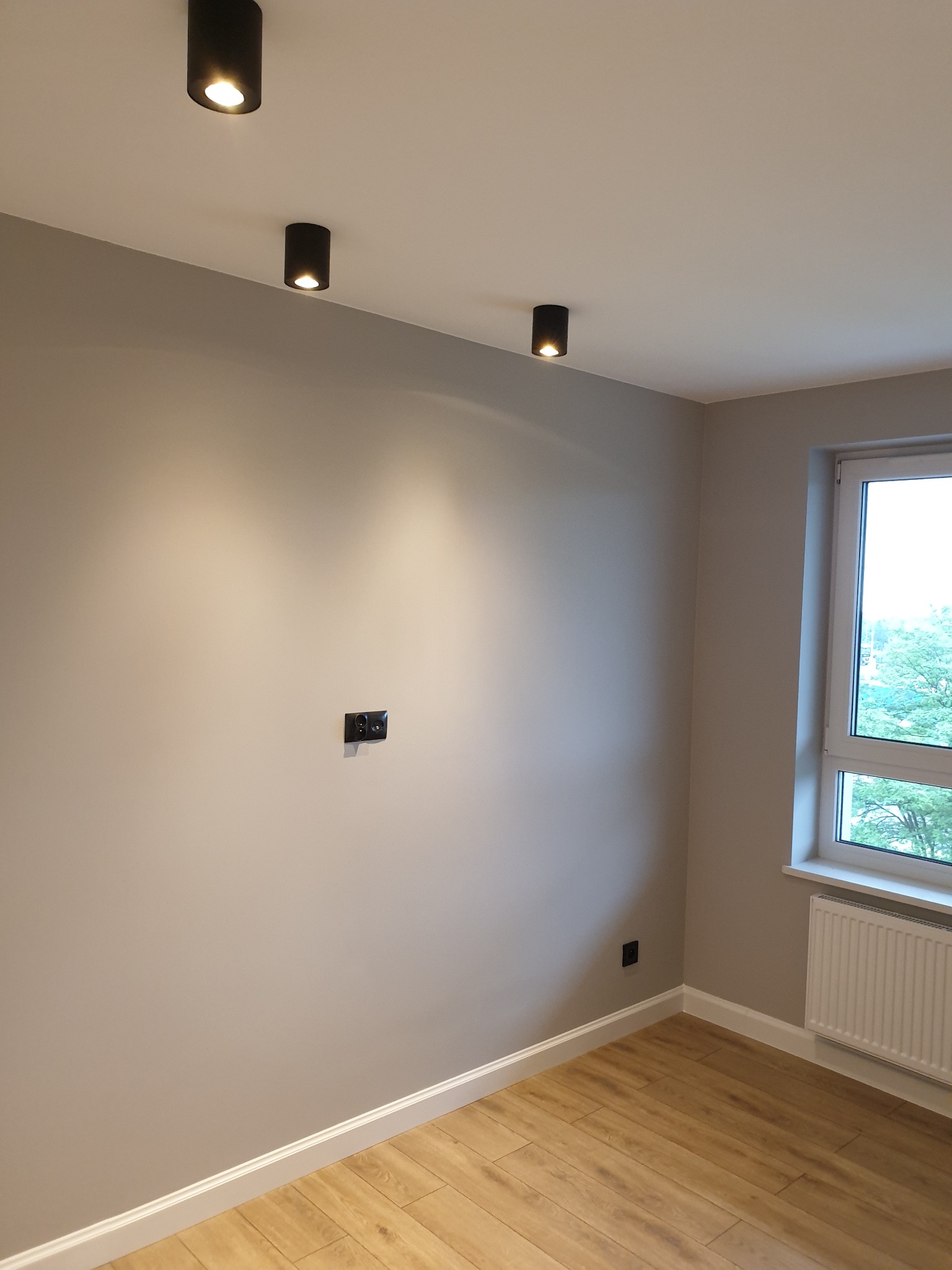 Pomalowane na szaro wnętrze pokoju z drewnianą podłogą, trzema czarnymi lampami sufitowymi, grzejnikiem pod oknem i podwójnym gniazdkiem elektrycznym w ścianie.