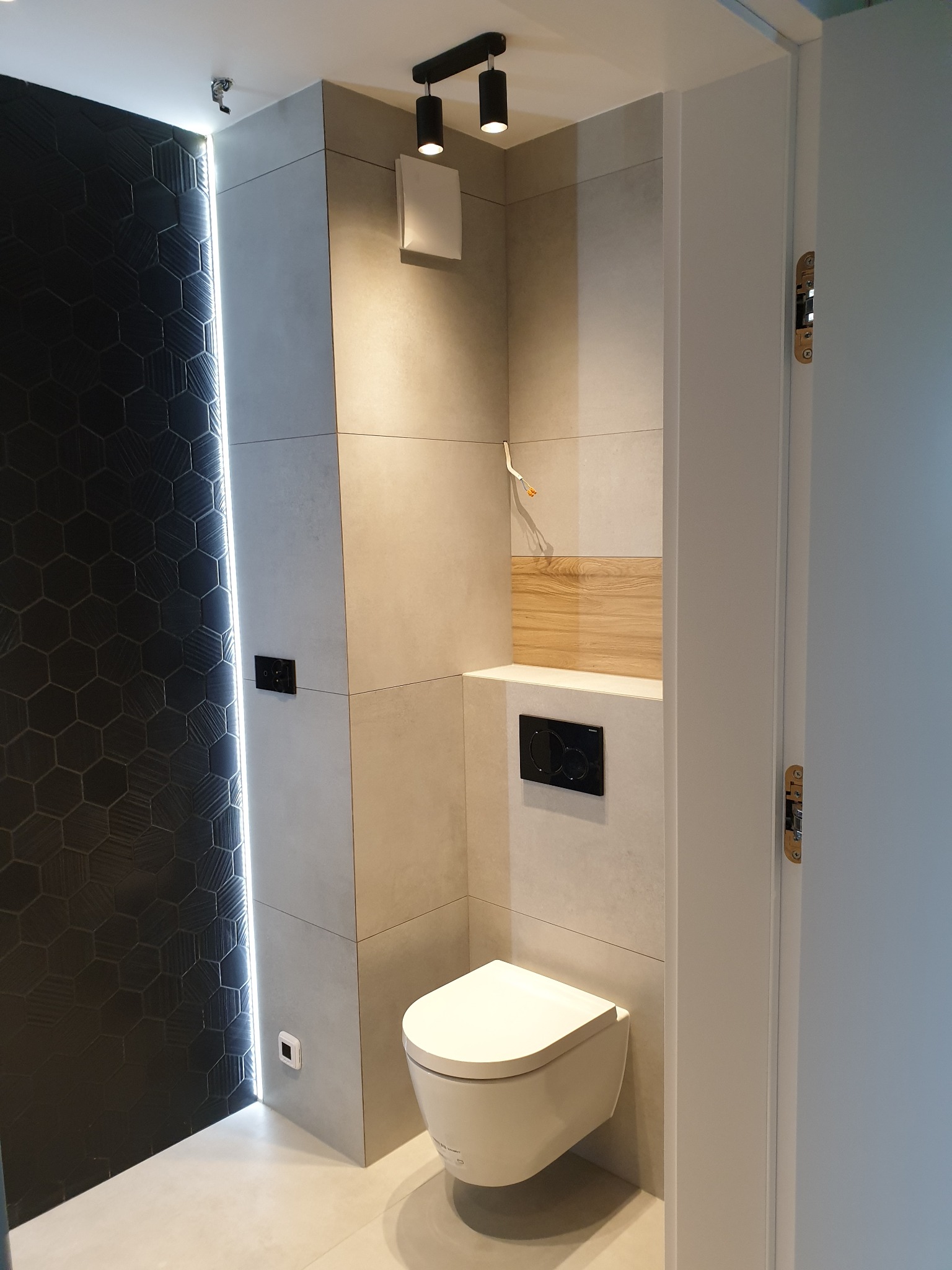Nowoczesna łazienka z czarną ścianą z heksagonalnych płytek z oświetleniem LED, zawieszona toaleta, szare płytki wielkoformatowe i drewniany akcent nad spłuczką.