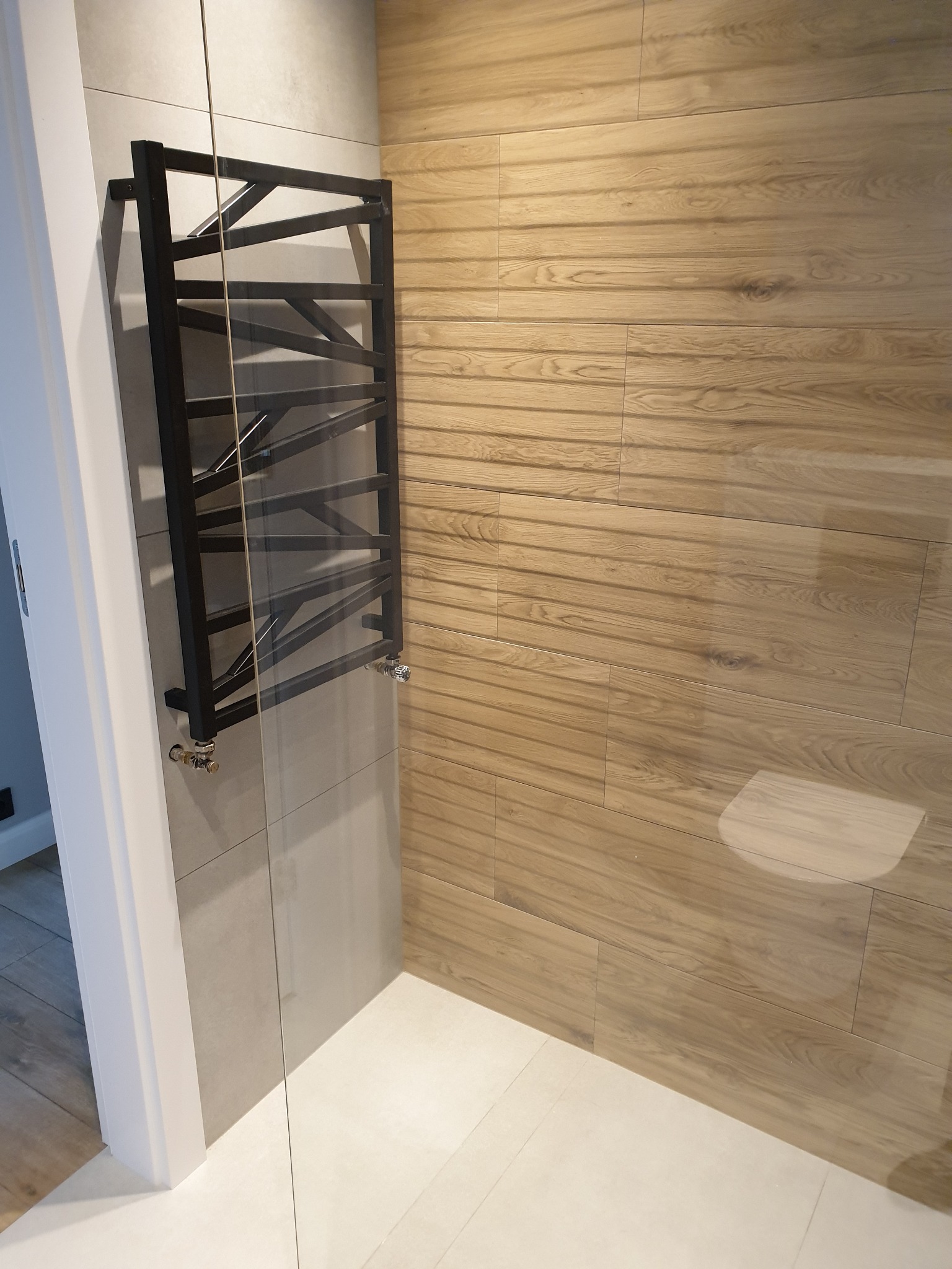 Nowoczesna łazienka z grzejnikiem drabinkowym w kolorze czarnym, drewnopodobnymi płytkami ściennymi i jasnymi płytkami podłogowymi, oddzielona szklaną ścianką.