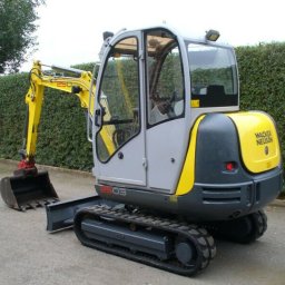 Wojtek - Żółto-szara mini koparka Wacker Neuson z łyżką, stojąca na betonowej powierzchni, z żywopłotem w tle.