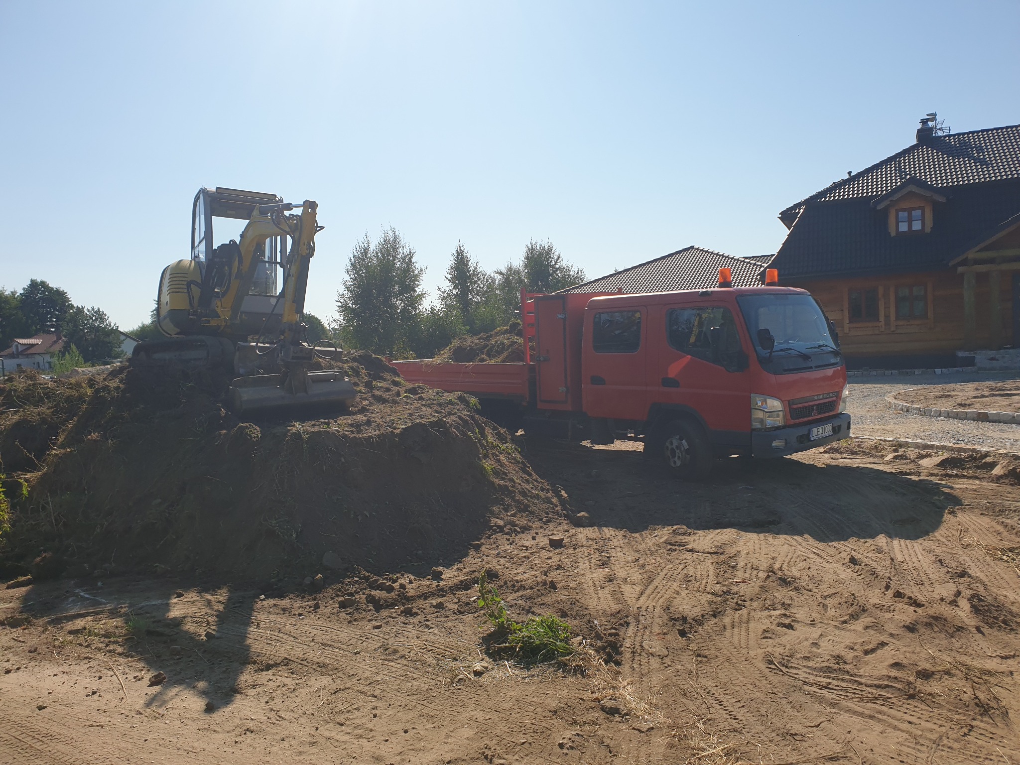 Żółta mini koparka na stercie ziemi obok czerwonej ciężarówki na tle drewnianego domu z ciemnym dachem w słoneczny dzień.