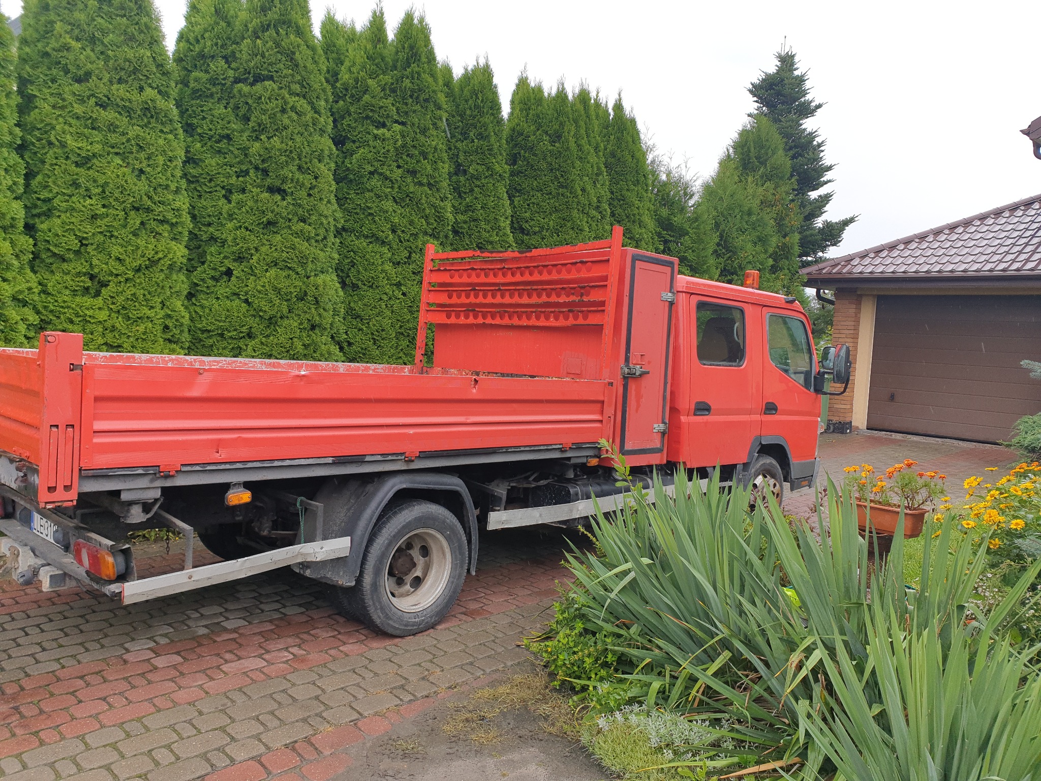 Czerwona wywrotka zaparkowana na posesji z kostki brukowej, tło stanowią wysokie tuje i dom z brązową bramą garażową.