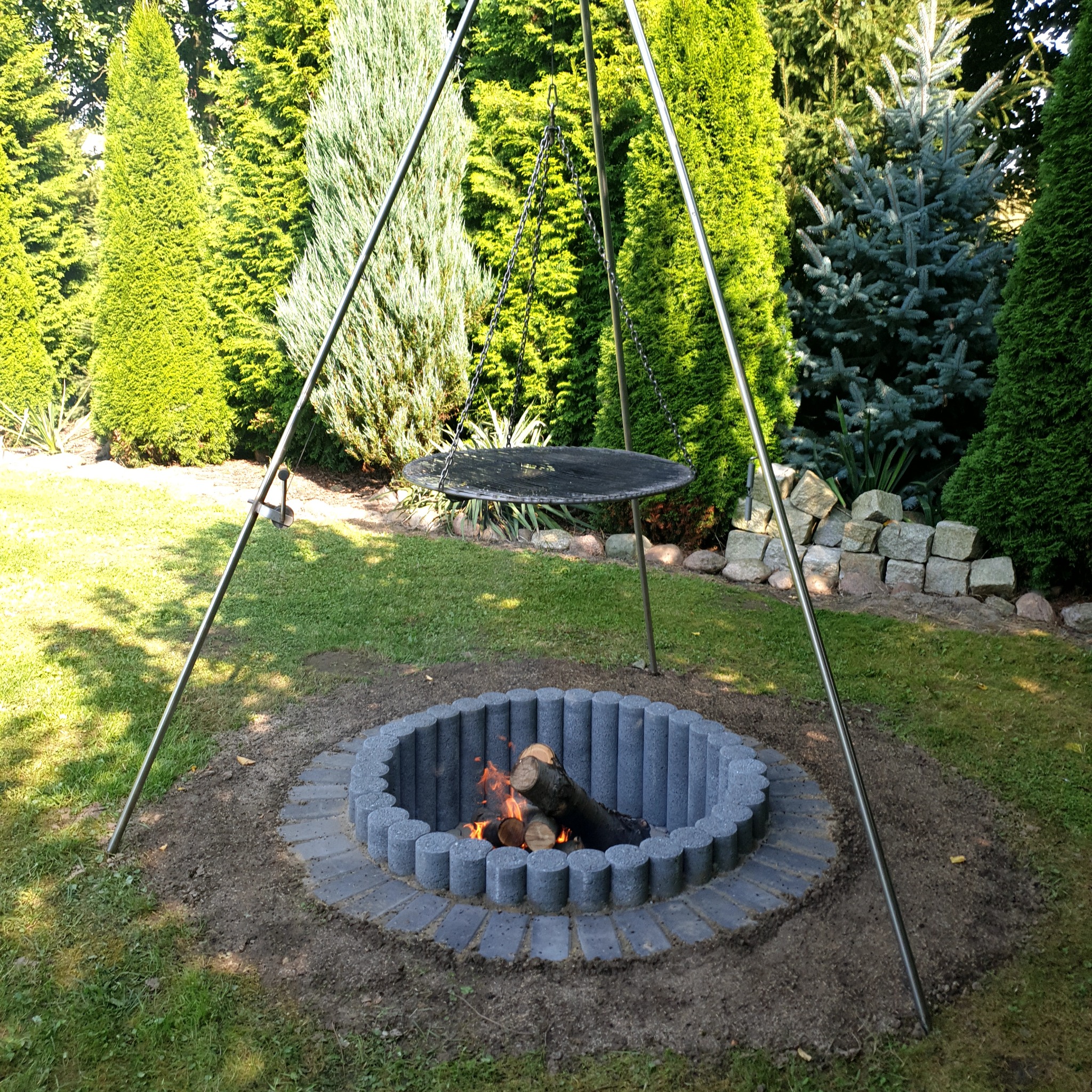 Okrągłe palenisko z szarej kostki betonowej, otoczone ziemią i trawą, z płonącym drewnem wewnątrz oraz metalowy trójnóg z rusztem nad paleniskiem, w tle zielone drzewa iglaste.