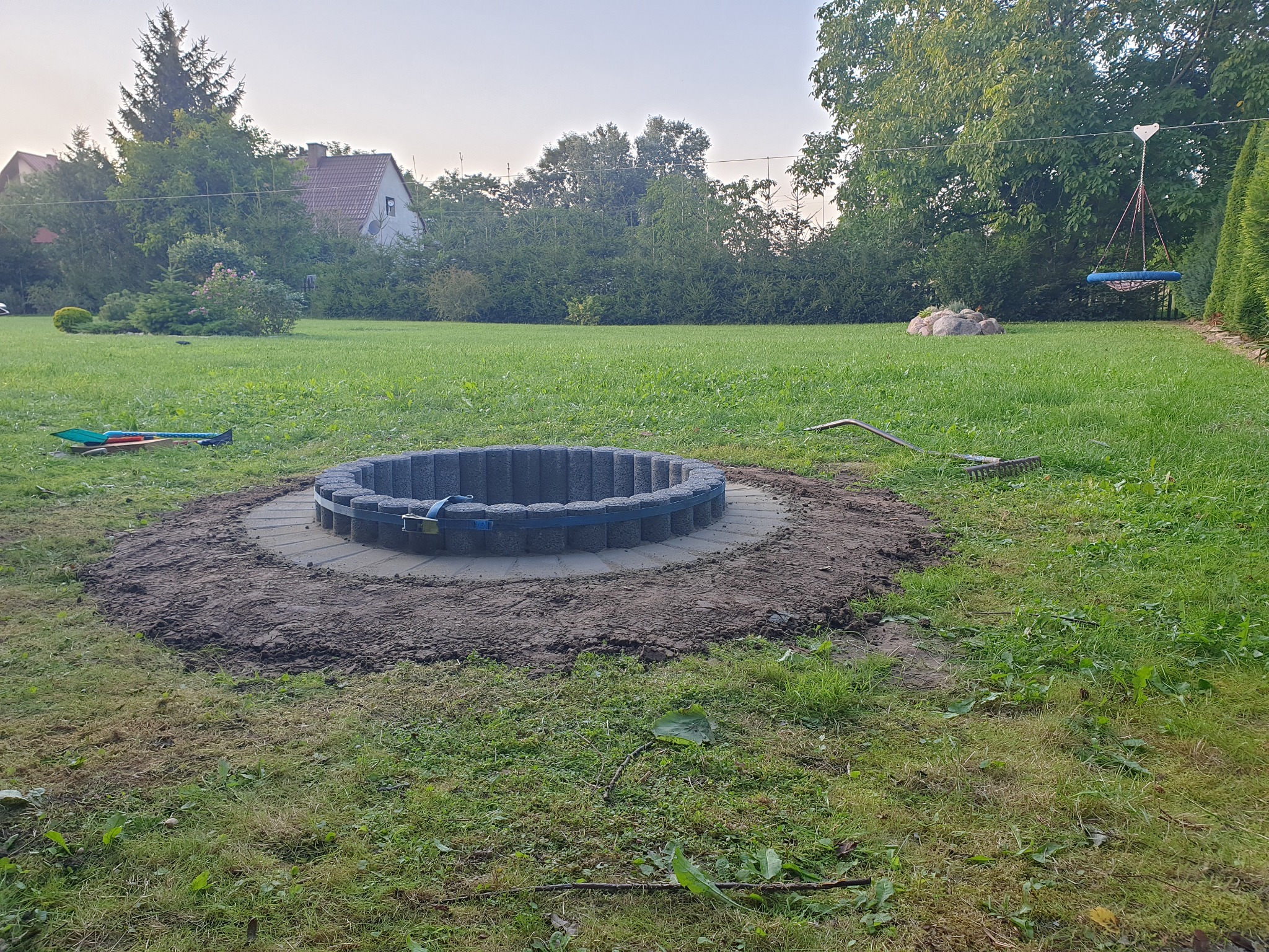 Okrągła konstrukcja z betonowych elementów, zabezpieczona niebieskim pasem, w trakcie budowy na trawniku, z widocznymi narzędziami ogrodniczymi w tle.