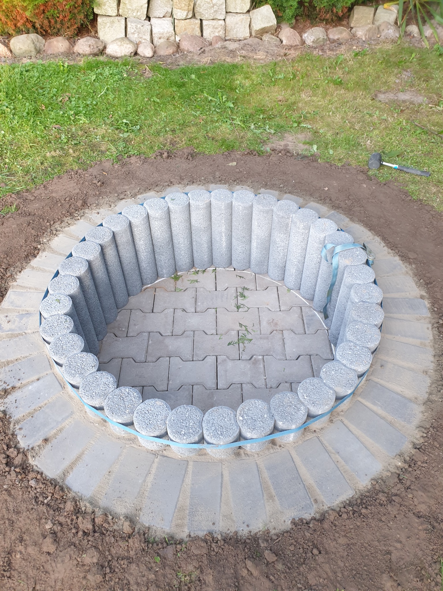 Okrągłe palisady z szarego betonu, tymczasowo spięte niebieską taśmą, tworzą obramowanie dla wnętrza wyłożonego szarą kostką; w tle widoczny gumowy młotek.