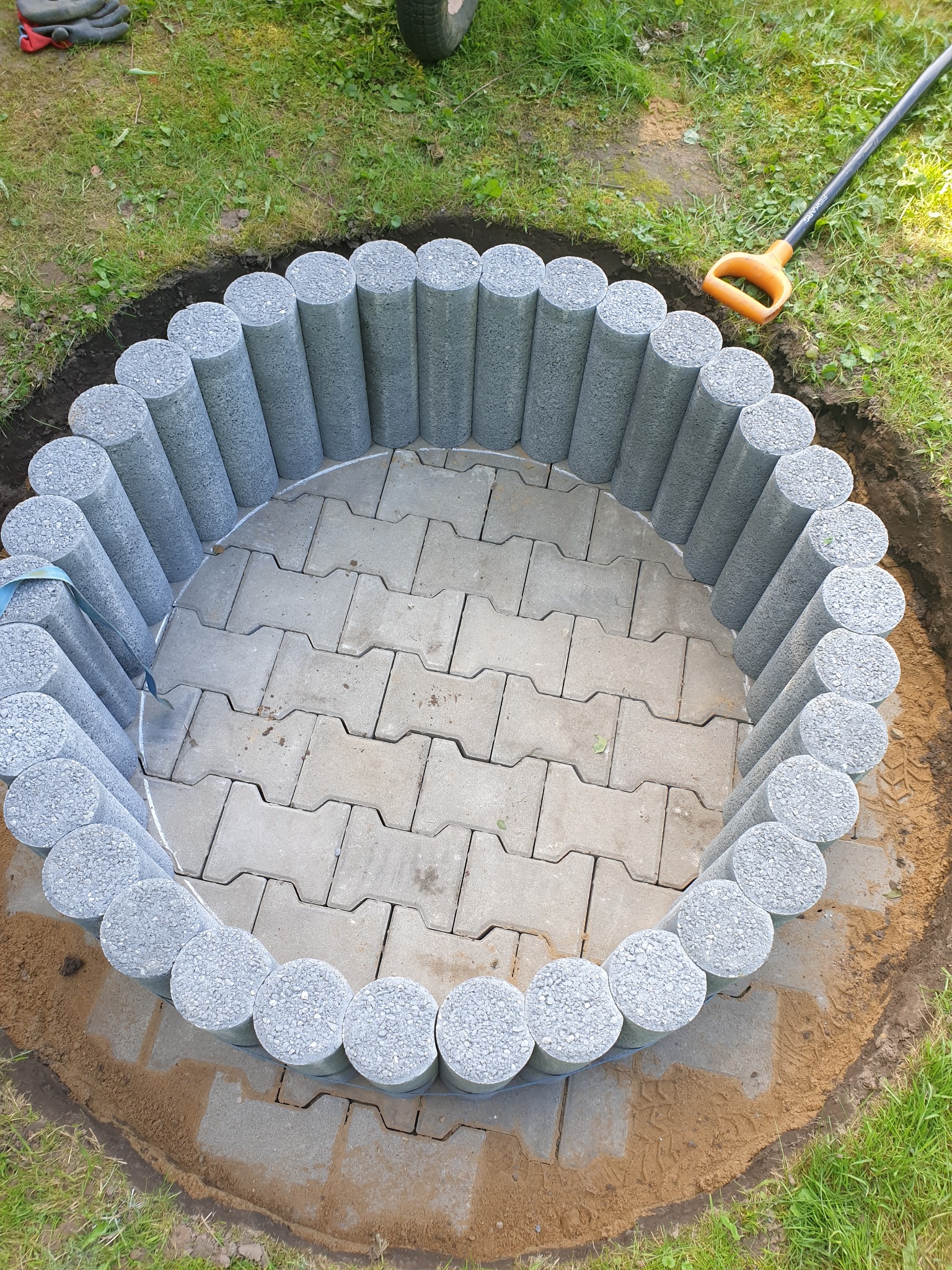 Okrągłe palisady z szarego betonu tworzą obrzeże wkoło ułożonej kostki brukowej, widoczna łopata i wykopana ziemia. Projekt w trakcie realizacji.