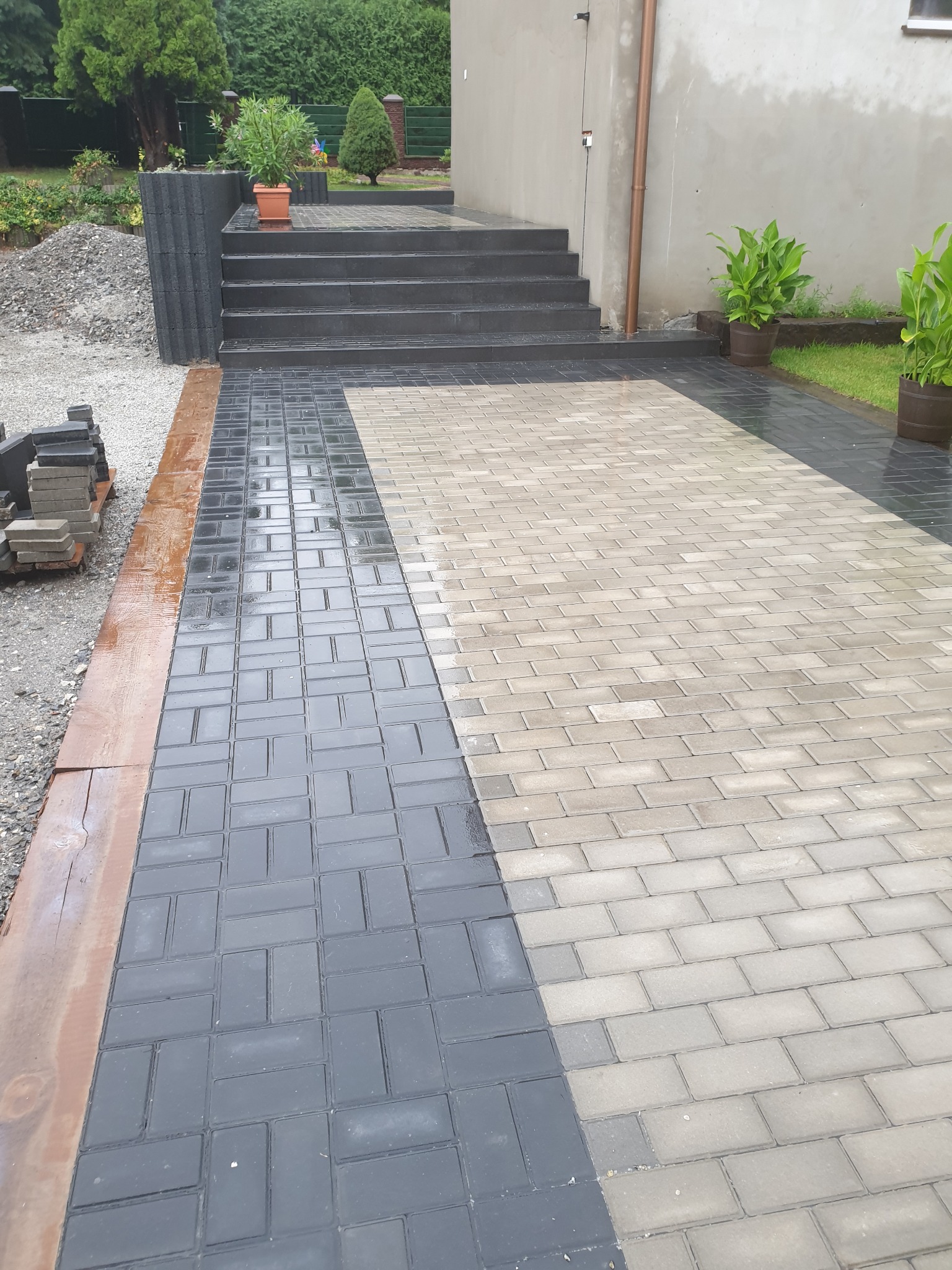 Ułożona kostka brukowa w dwóch odcieniach szarości, tworząca wzór na podjeździe, z widocznymi schodami i roślinami w donicach w tle, oraz stosem kostki po lewej stronie.