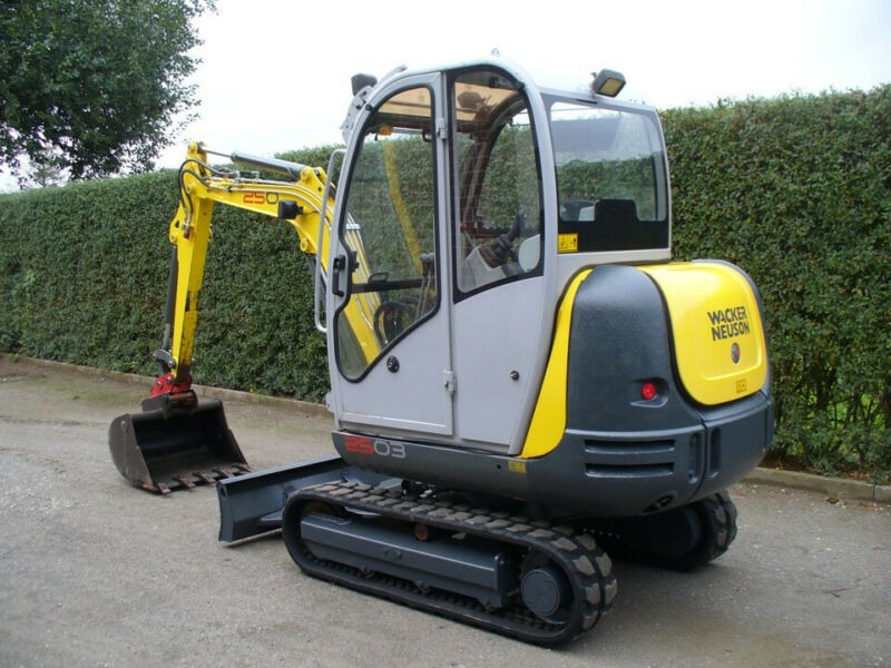 Żółto-szara mini koparka Wacker Neuson z łyżką, stojąca na betonowej powierzchni, z żywopłotem w tle.