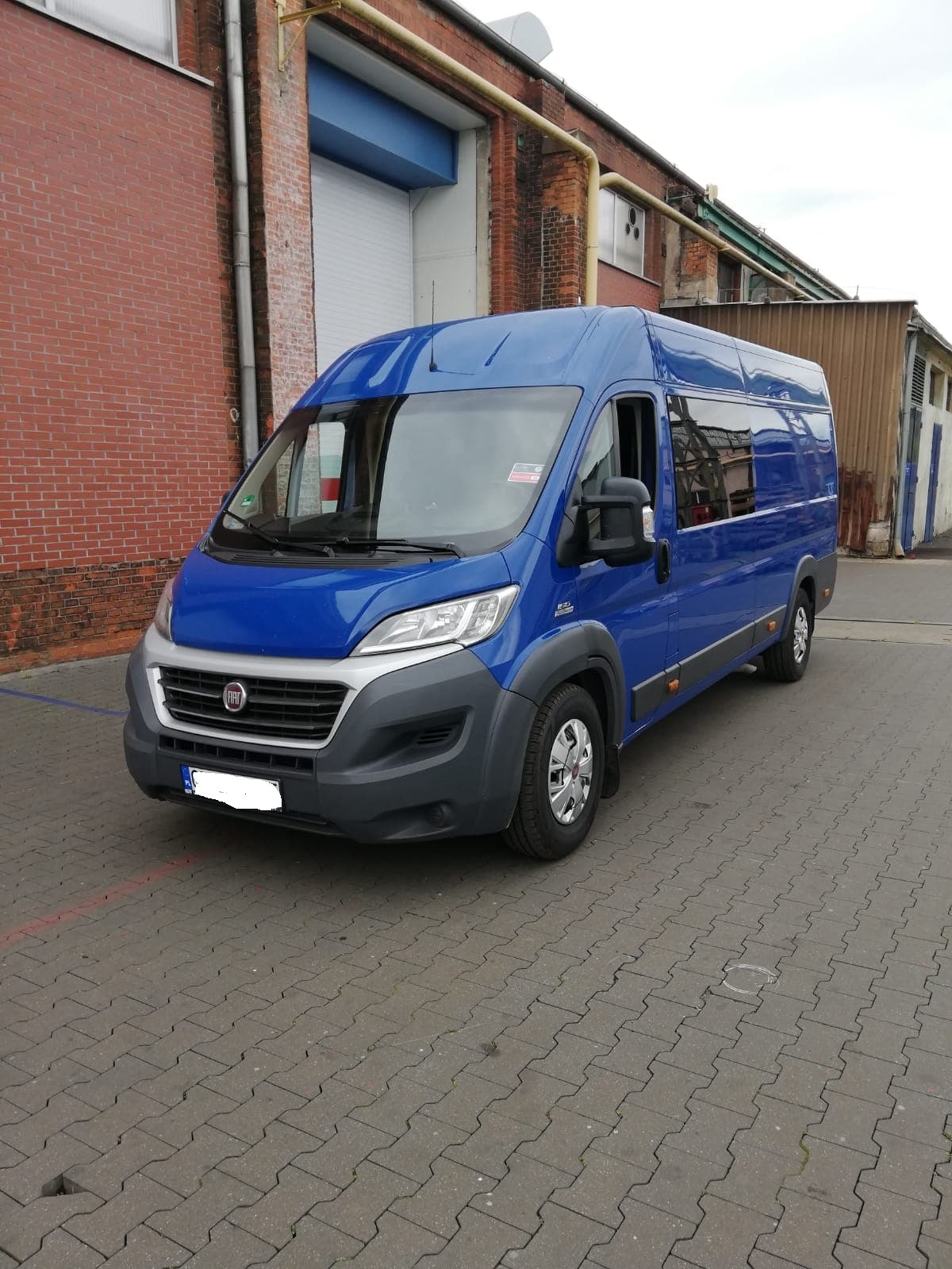 Niebieski bus Fiat Ducato zaparkowany na szarej kostce brukowej przed ceglanym budynkiem z bramą rolowaną.