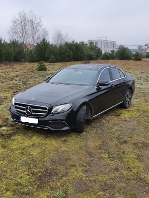 Czarny Mercedes-Benz E-Klasa na tle jesiennego krajobrazu z widocznymi drzewami iglastymi i budynkiem w oddali, uchwycony z niskiego kąta.