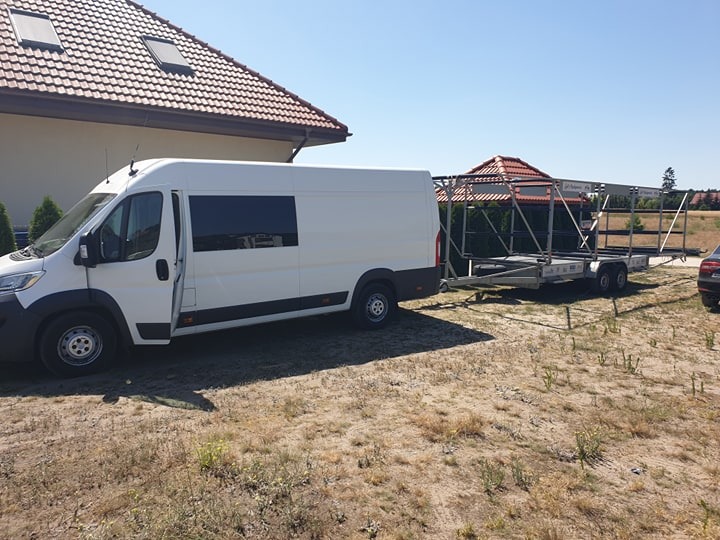 Biały bus z otwartymi drzwiami i przyczepą z metalową konstrukcją na tle domu z czerwoną dachówką, w letni dzień.