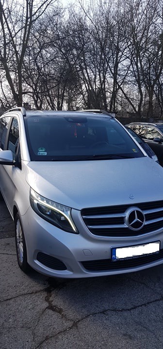 Srebrny Mercedes V-klasy zaparkowany na zewnątrz, widok z przodu, z widocznymi światłami LED i drzewami w tle.