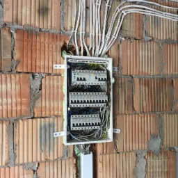 Rozdzielnica elektryczna z zabezpieczeniami zamontowana na ścianie z cegły, przewody w peszlach mocowane klamrami, pianka montażowa widoczna wokół peszli.