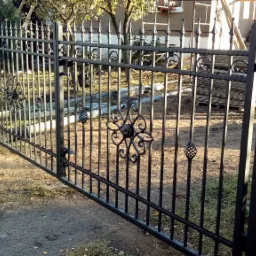 Czarna brama wjazdowa z metaloplastyki, zdobiona motywem kwiatowym, widoczna na tle posesji z budynkiem i drzewami, z fragmentem siatki ogrodzeniowej po prawej stronie.