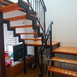 Metalowe schody z drewnianymi stopniami i ozdobnymi kutymi balustradami, zabezpieczone bramką dla dziecka, widok z dołu na jasnym tle ściany.