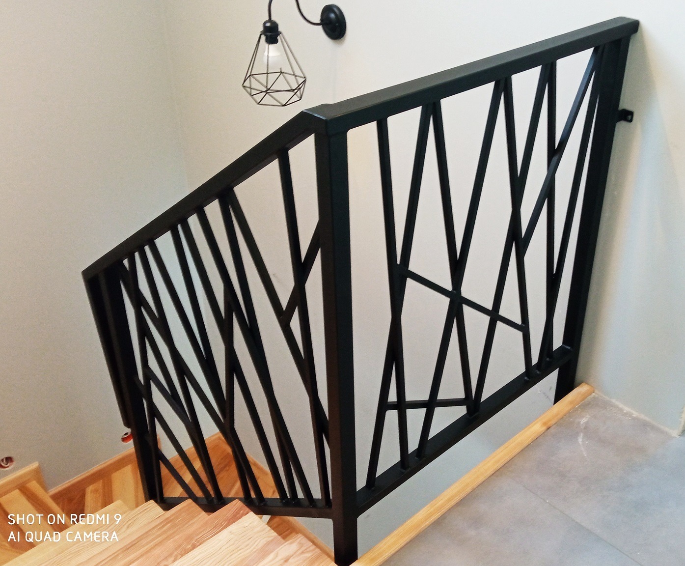 Nowoczesna, czarna balustrada metalowa schodów wewnętrznych z geometrycznym wzorem, oświetlona lampą ścienną w stylu industrialnym.