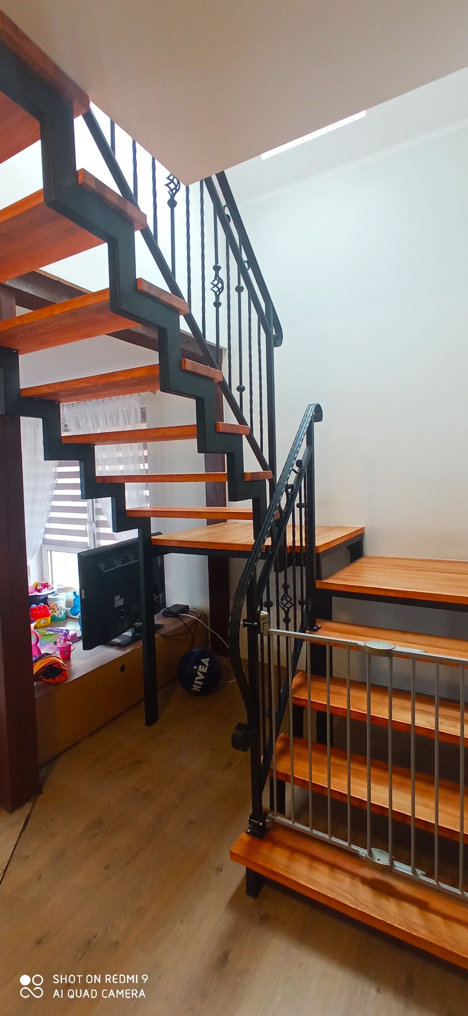 Metalowe schody z drewnianymi stopniami i ozdobnymi kutymi balustradami, zabezpieczone bramką dla dziecka, widok z dołu na jasnym tle ściany.