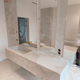 Jakub Matuszny JACOBS - Wykończona łazienka z podwieszaną szafką umywalkową z blatem imitującym marmur, pionowymi lustrami i płytkami w tym samym stylu. Widoczne gniazdka elektryczne i złota bateria.