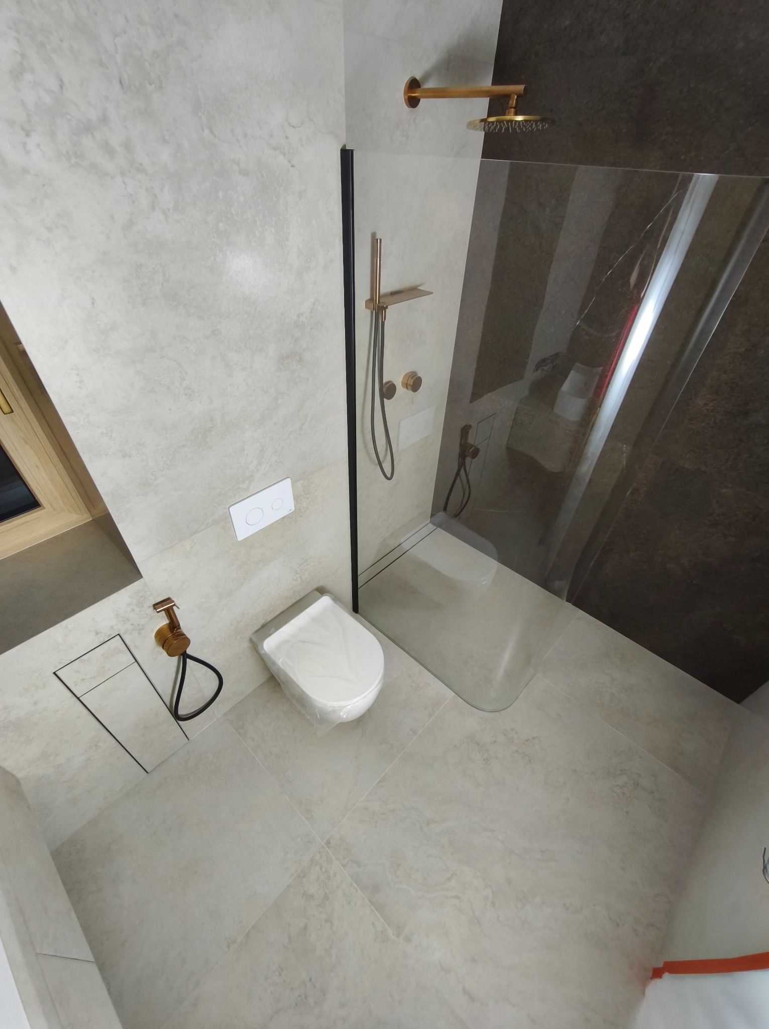 Jasna łazienka z kabiną typu walk-in, wykończona płytkami imitującymi kamień. Uwagę przykuwają złote detale armatury i czarne ramy kabiny.