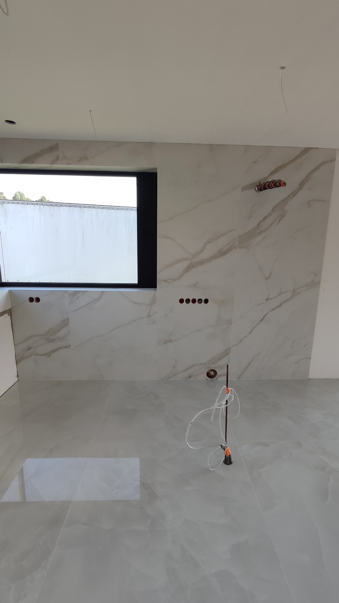 Wykończona podłoga i ściany w jasnych płytkach imitujących marmur. Widoczne otwory pod gniazdka i wystające kable. Okno z widokiem na zewnątrz.