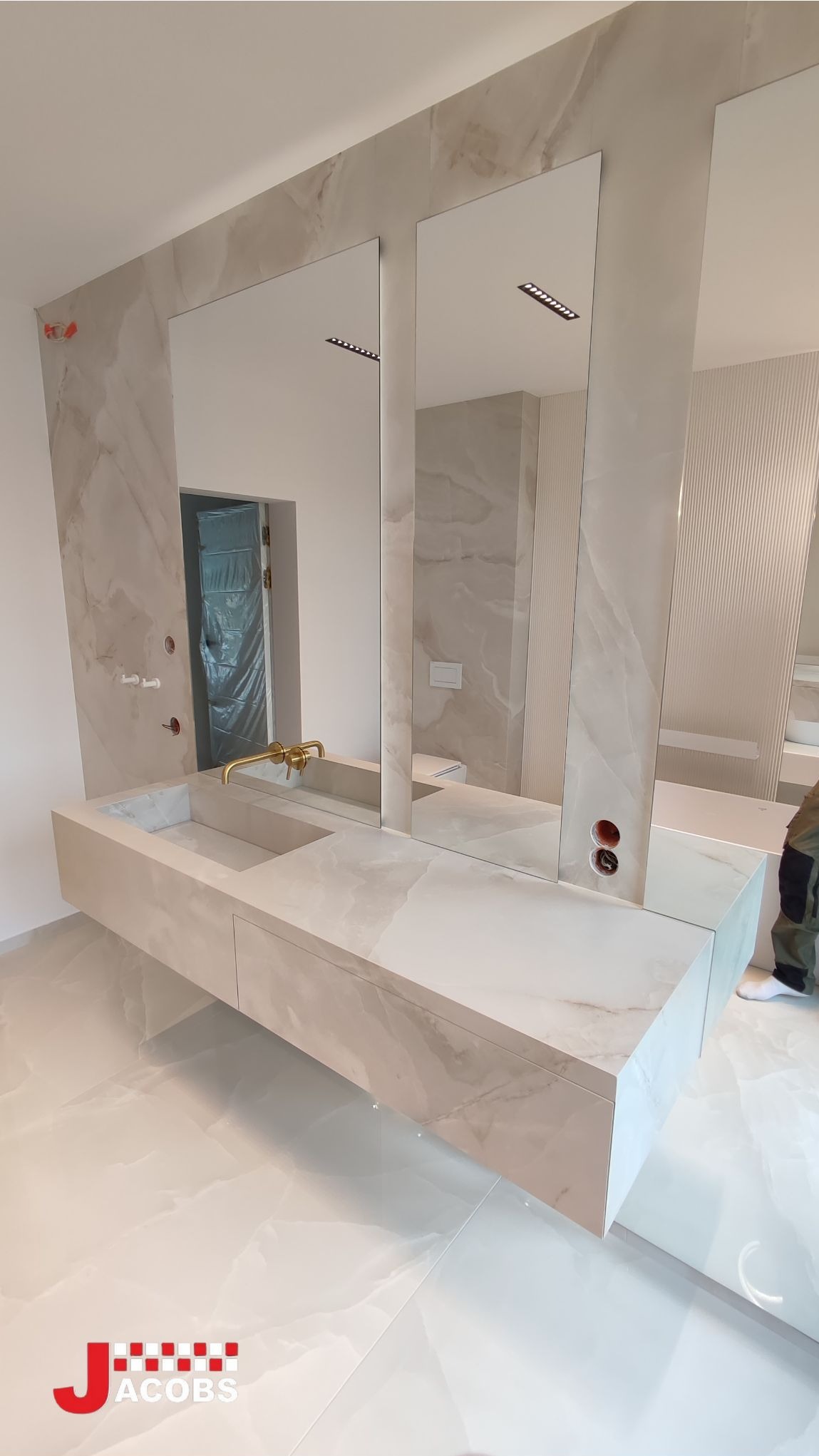 Wykończona łazienka z podwieszaną szafką umywalkową z blatem imitującym marmur, pionowymi lustrami i płytkami w tym samym stylu. Widoczne gniazdka elektryczne i złota bateria.