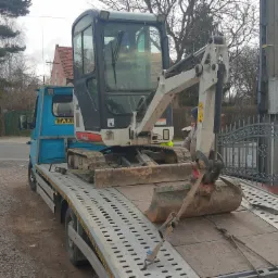 Niebieska laweta z napisem 'TAXI' przewozi brudną, biało-szarą mini-koparkę Bobcat na platformie zabezpieczonej pasami transportowymi. Koparka z łyżką skierowaną w stronę kabiny lawety.