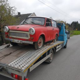 Auto Serwis Krystan Sendor - Czerwony Trabant z widocznymi śladami użytkowania, zabezpieczony pasami na ażurowej platformie niebieskiej lawety, zaparkowanej na szarej asfaltowej drodze.