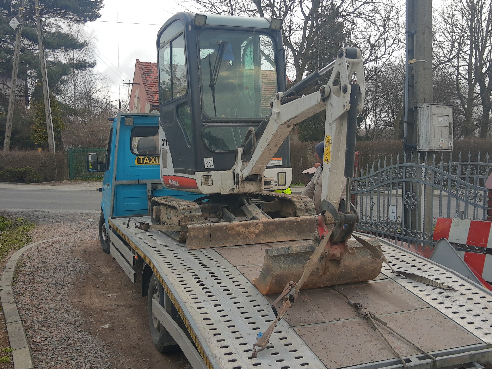 Niebieska laweta z napisem 'TAXI' przewozi brudną, biało-szarą mini-koparkę Bobcat na platformie zabezpieczonej pasami transportowymi. Koparka z łyżką skierowaną w stronę kabiny lawety.