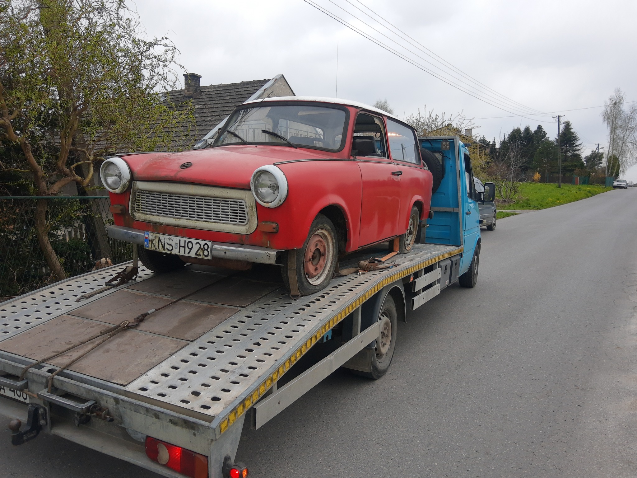 Czerwony Trabant z widocznymi śladami użytkowania, zabezpieczony pasami na ażurowej platformie niebieskiej lawety, zaparkowanej na szarej asfaltowej drodze.